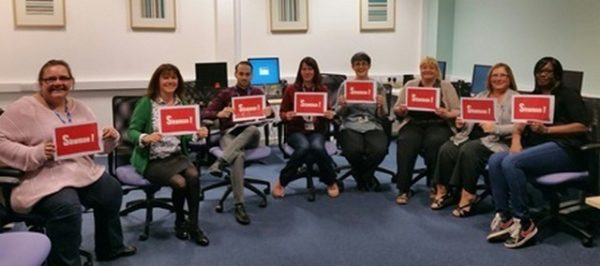 8 people sitting in a semi circle holding up signs saying "shwmae"