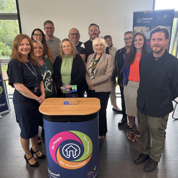 On the far right, Rob Milligan from Tai Pawb stands in front of Cartrefi Conwy's banner. To the left of him are 11 staff/board members from Cartrefi Conwy. In front of them all is the QED glass trophy on a plinth.
