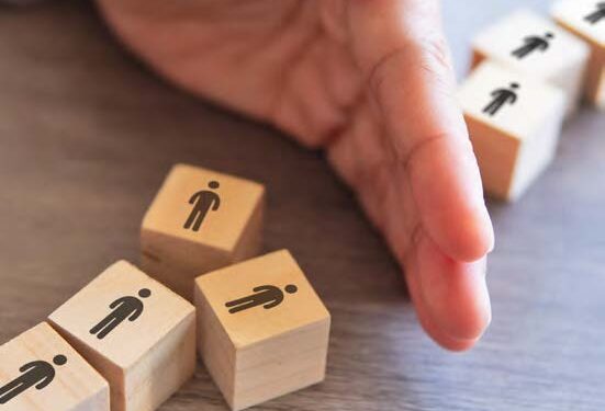 Image of a hand inbetween two sets of wooden blocks with sillouette images of people on them. To depict a block to some people in the queue