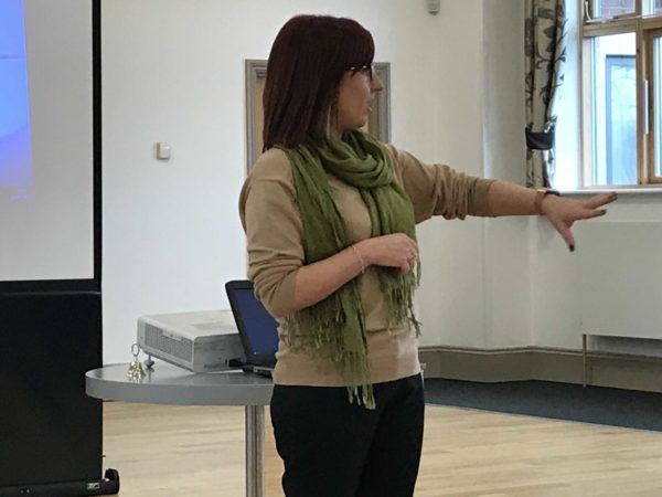 A woman presenting and gesturing with her arms in front of a laptop and projector