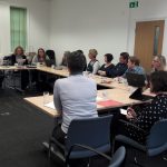 Group of people sat around a U-shaped conference table listening to an unseen presenter