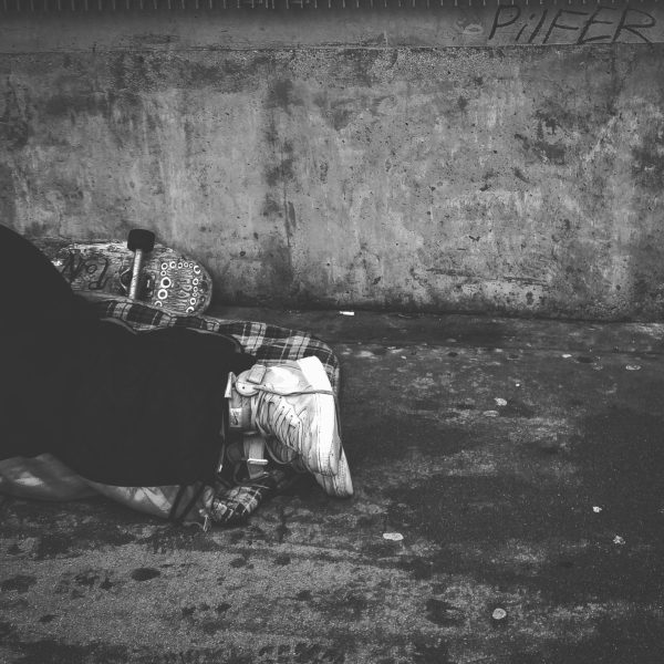 Black and white picture of someone's legs sleeping on the streets. They are wearing dirty high top trainers and have a skateboard behind them