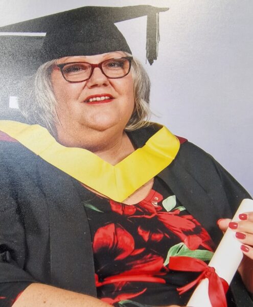 Photo of Jill Wadley. Jill is wearing a cap and gown at her graduation, with a red dress and a diploma in hand. Jill is a white women with grey hair and glasses and is in. a wheelchair.
