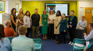 Group picture of Tai Pawb and Valleys to Coast staff stood in front of a presentation screen, holding the glass QED award. There are other staff in seats watching on.